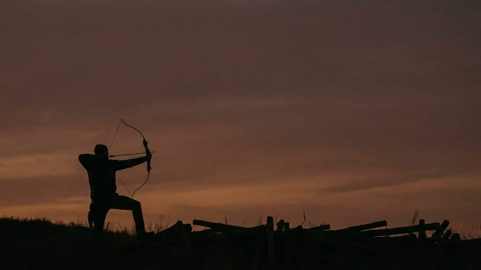 An archer at full draw silhouetted against a dusk sky above a timbered ridge
